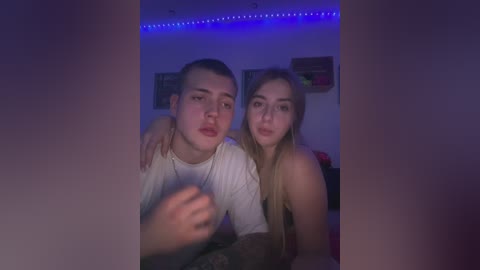 Media: Video of two young Caucasian teenagers, a boy and a girl, sitting close together in a dimly lit room with blue LED lights. The boy, wearing a white shirt, has short hair; the girl, with long blonde hair, is topless.