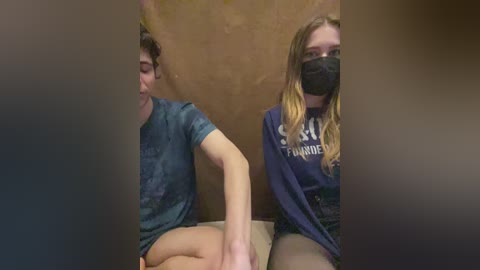 Video of two young women, one with curly hair, one with long blonde hair, both in masks, sitting on a couch with a beige cushion, in a dimly lit room.