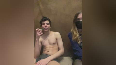Video: Young, shirtless, fair-skinned man with dark hair, wearing gray shorts, holding cigarette. Next to him, masked, fair-skinned woman with long blonde hair, wearing dark shirt. Brown, textured background.