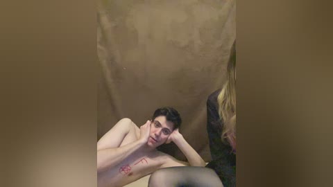 A video captures a shirtless young man with short dark hair, lying on a beige sheet, his hand resting on his face. Another person, partially visible, with blonde hair and a dark patterned garment, is next to him.