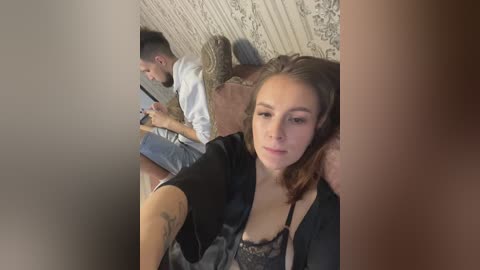 Media: Video of a young woman with light skin and long brown hair, wearing a black lace bra, taking a selfie. Background shows a man with dark hair and a beard, seated on a couch.