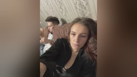 Media: Video of a woman with light brown hair in a black robe, lying next to a man with short dark hair, both on a bed with patterned wallpaper, looking tired.