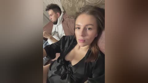 Media: A video of a young woman with light skin and straight brown hair, wearing a black satin blouse, sitting on a train, next to a man in a hoodie, both looking at their phones.