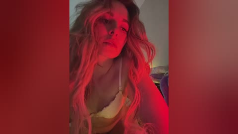 A video of a woman with long, wavy blonde hair, wearing a yellow lace bra, bathed in red light, leaning forward with a pensive expression.