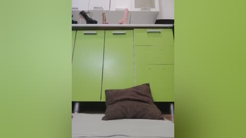 Video of a modern kitchen with lime-green cabinets, a grey countertop, and a white sink. A dark brown cushion sits on a white stool.