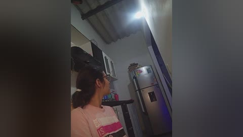 Video of a woman with dark hair in a ponytail, wearing glasses and a pink shirt, standing in a dimly lit, narrow hallway with a fridge and a ceiling light.