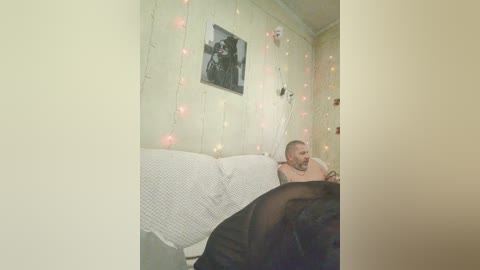 A video of a bearded man in a white shirt, sitting on a bed with white sheets, in a cozy room adorned with string lights and a framed black-and-white photo on the wall.