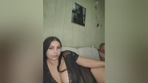 Video of a light-skinned woman with long black hair, wearing a black lace nightgown, sitting on a white couch in a dimly lit room with green walls and a framed photo.