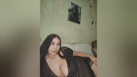 Video of a young Latina woman with long black hair, large breasts, and dark eyes, wearing a sheer black lace lingerie top, lying on a white couch in a cozy, softly lit room with string lights and a framed black-and-white photo on the wall.