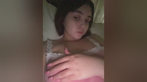 Media: Video of a young woman with fair skin and brown hair, lying on her back in bed, wearing a white lace bra, her hand resting on her stomach, with dimly lit, neutral-toned walls in the background.