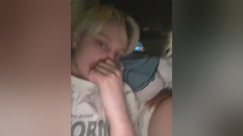 Media: Video of a young blonde girl with fair skin, lying on a bed, covering her mouth with her hand, wearing a white T-shirt with black text. The background is dimly lit with blurred objects.