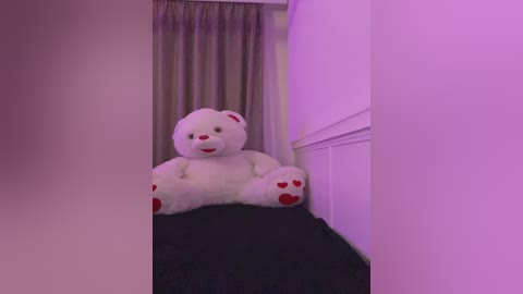 Video of a plush white teddy bear with red paw pads, sitting on a black cushion in a dimly lit room with purple lighting, partially visible through a doorway.
