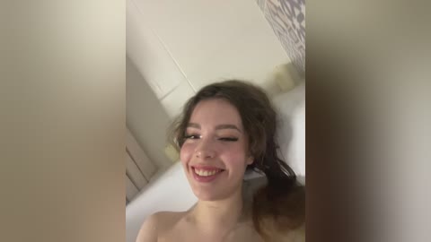 Video of a young, smiling woman with long brown hair, fair skin, and no visible clothing, standing in a modern bathroom with white tiles and a bathtub.