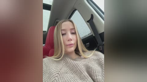 A video of a young woman with long blonde hair, wearing a beige sweater, seated in a car with red seats.