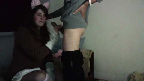 Media: Video of a young woman with light skin, long brown hair, and pink cat ears, sitting on a bed, holding a dildo, wearing a brown sweater and white shorts, next to a man wearing a gray shirt and black pants.