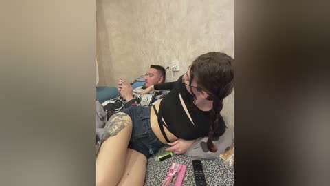 Media: Video of a young woman with a tattooed thigh, wearing a black crop top and denim shorts, sitting next to a man on a bed, both holding phones. Background: beige wall, cluttered bedside table.