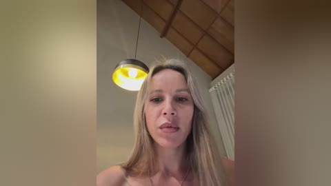 Media: Video of a blonde woman with fair skin, medium build, and small breasts, standing indoors with a wooden ceiling, a yellow pendant light, and a radiator in the background.
