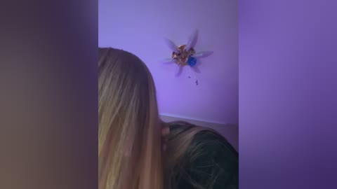 Media: Video of a blonde woman with long hair, wearing a dark green top, leaning against a wall with a purple light, gazing at a decorative, fairy-themed wall sconce.