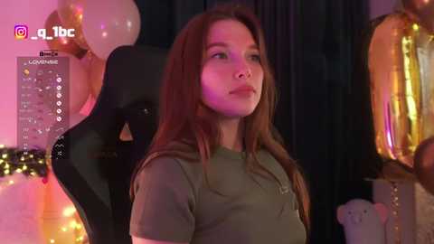 Video of a young woman with long brown hair, wearing a green top, seated in a black gaming chair. Background includes gold and white balloons, dim lighting, and social media icons.