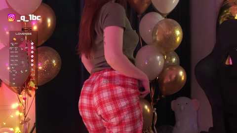 Video of a curvy woman with long brown hair, wearing a tight, gray top and red plaid skirt, holding gold balloons. Background includes dark walls, string lights, and a white bear toy.