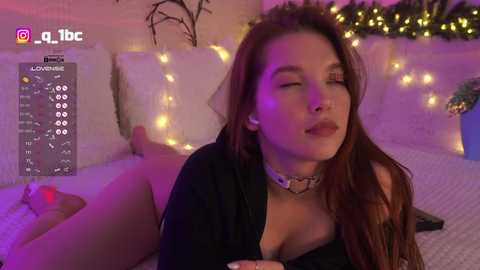 A video of a smiling woman with long brown hair and a black choker, lying on a bed with white pillows and a cozy, dimly-lit room with fairy lights.