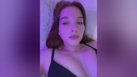 Video of a young woman with light skin and long auburn hair, wearing a black spaghetti-strap top, lying on a bed with soft, white bedding. The image is softly lit with purple hues, creating a serene, intimate atmosphere.