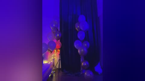 Video of a dimly-lit room with a black curtain backdrop, featuring clusters of gold and white balloons and a string of fairy lights on the left.