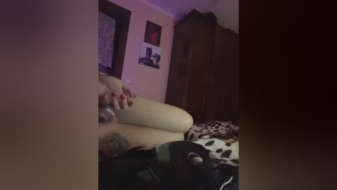 Video of a dimly lit bedroom featuring a nude person with fair skin, lying on a bed with leopard-print sheets, surrounded by personal items and a wardrobe.