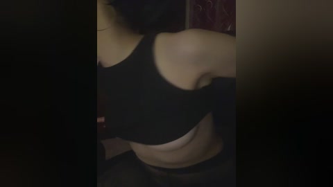 A dimly lit video of a woman with medium skin tone, wearing a black sports bra and black pants, partially unbuttoned. Her head is slightly out of frame, revealing her shoulder and upper chest.