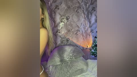 Media: Video of a nude blonde woman with long hair, sitting on a bed with a textured silver blanket. The room features a mural of tree silhouettes and a lit Christmas tree in the background.