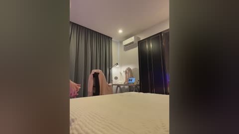 Video of a dimly lit, modern hotel room with beige walls, a white bed, dark curtains, an air conditioner, and a desk with a laptop.