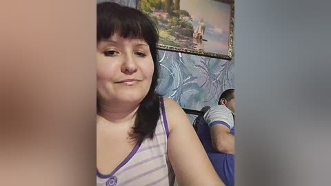 Video of a plus-sized woman with dark hair and bangs, wearing a striped tank top, standing in a room with a tropical painting and a man in blue shorts.