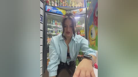 Media: Video of a young woman with dark hair, wearing a light blue button-up shirt, bending over to reach for a bottle in a colorful, cluttered convenience store fridge.