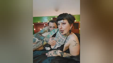 Video of a tattooed woman with short dark hair, wearing black lingerie, lying on a bed with a man partially visible behind her.