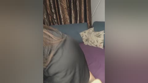 Video of a person in a gray hoodie, partially obscured by a purple pillow, standing beside a bed with a floral-patterned pillow and brown, textured curtains in the background.