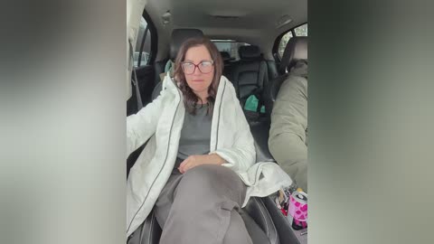 Media: Video of a middle-aged woman with glasses and shoulder-length brown hair, wearing a white jacket over a grey sweater, seated in the back seat of a car, surrounded by bags and other items.