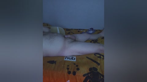 Video of a man lying on a bed with a yellow, patterned duvet featuring cartoon characters. His bare, hairy legs are visible, and his hand rests on his abdomen. The background is dimly lit, with a hint of a white wall and a blue object.