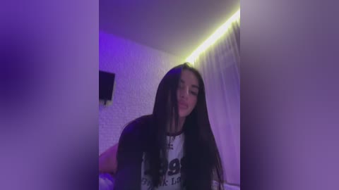 Media: Video of a young woman with long, straight black hair, wearing a black t-shirt with white text, smiling in a dimly lit, purple-lit bedroom.