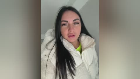 Video of a young woman with long black hair, wearing a white puffy jacket and a lime green shirt, standing against a plain white background.