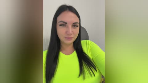 Video of a young woman with long black hair, wearing a bright neon green t-shirt, seated indoors against a plain, light-colored wall. The image is slightly blurred, with a pink and yellow gradient filter applied.