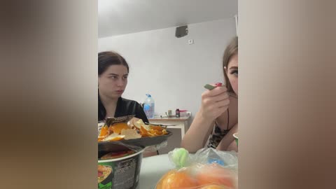 Media: Video of two women in a kitchen; one with dark hair, black shirt, and orange food, the other with blonde hair, green shirt, eating. Background shows a light wall and counter with water bottle and other items.