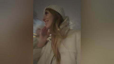 Media: A video of a smiling woman with long, wavy blonde hair, wearing a beige beanie and coat, captured from a low angle. The background is blurred, suggesting an indoor setting with soft lighting.