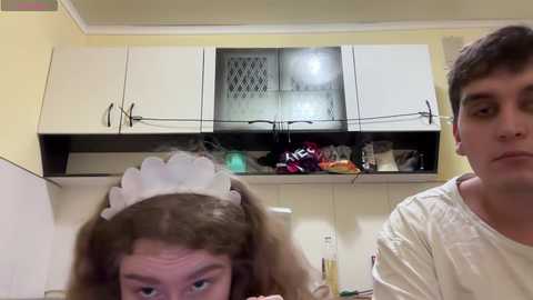 Media: Video of a young woman with curly brown hair wearing a white maid headband, smiling widely, and a young man with short brown hair in a white shirt, both in a kitchen with white cabinets and a black fridge.