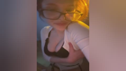 Media: Video of a fair-skinned woman with short, light brown hair, wearing black-rimmed glasses and a black bra, leaning against a white wall in a dimly lit room.