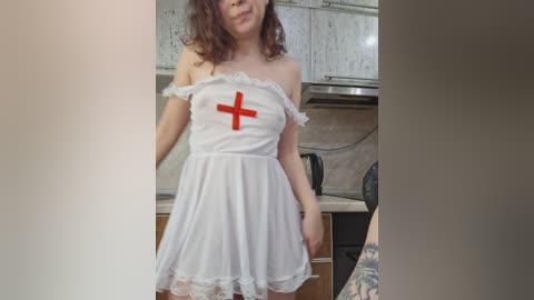 Media: Video of a young, light-skinned woman with curly brown hair, wearing a white, off-the-shoulder nurse dress with a red cross, standing in a modern kitchen.