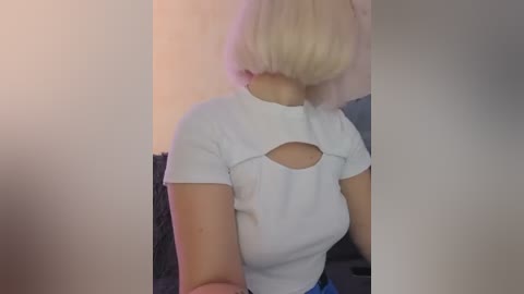 Video of a woman with a short, platinum blonde bob haircut, wearing a white, short-sleeved blouse with a cut-out design on the back. She stands in a dimly lit room with soft, warm lighting.