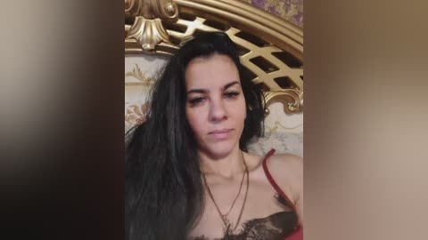 A video of a young woman with long, black hair, wearing a red lace bra and gold necklace, against a patterned wall and ornate gold headboard.