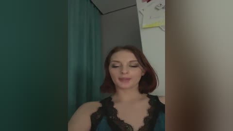 Media: A video of a Caucasian woman with shoulder-length auburn hair and light skin, wearing a black lace camisole, standing in a dimly lit room with teal and beige walls.