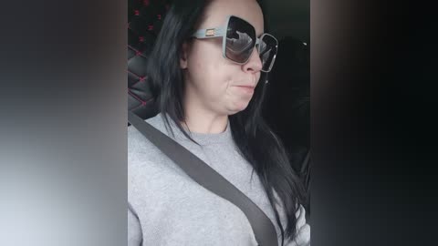 Video of a middle-aged woman with long black hair, wearing large, reflective sunglasses, a gray sweater, and a shoulder strap, seated in a car.