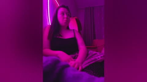 Video of a plus-size woman with long dark hair, wearing a black spaghetti strap top, sitting on a bed with a pink light illuminating the room.